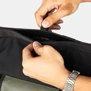 Manos abriendo un bolsillo interior con cremallera de la mochila táctica negra.